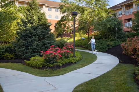 A woman walks a small white dog along a curved pathway through a landscaped courtyard beside a multi-story senior living building.