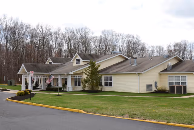 Exterior view of a single-story yellow senior living facility building with a covered entrance, an American flag, and a surrounding lawn with trees in the background.