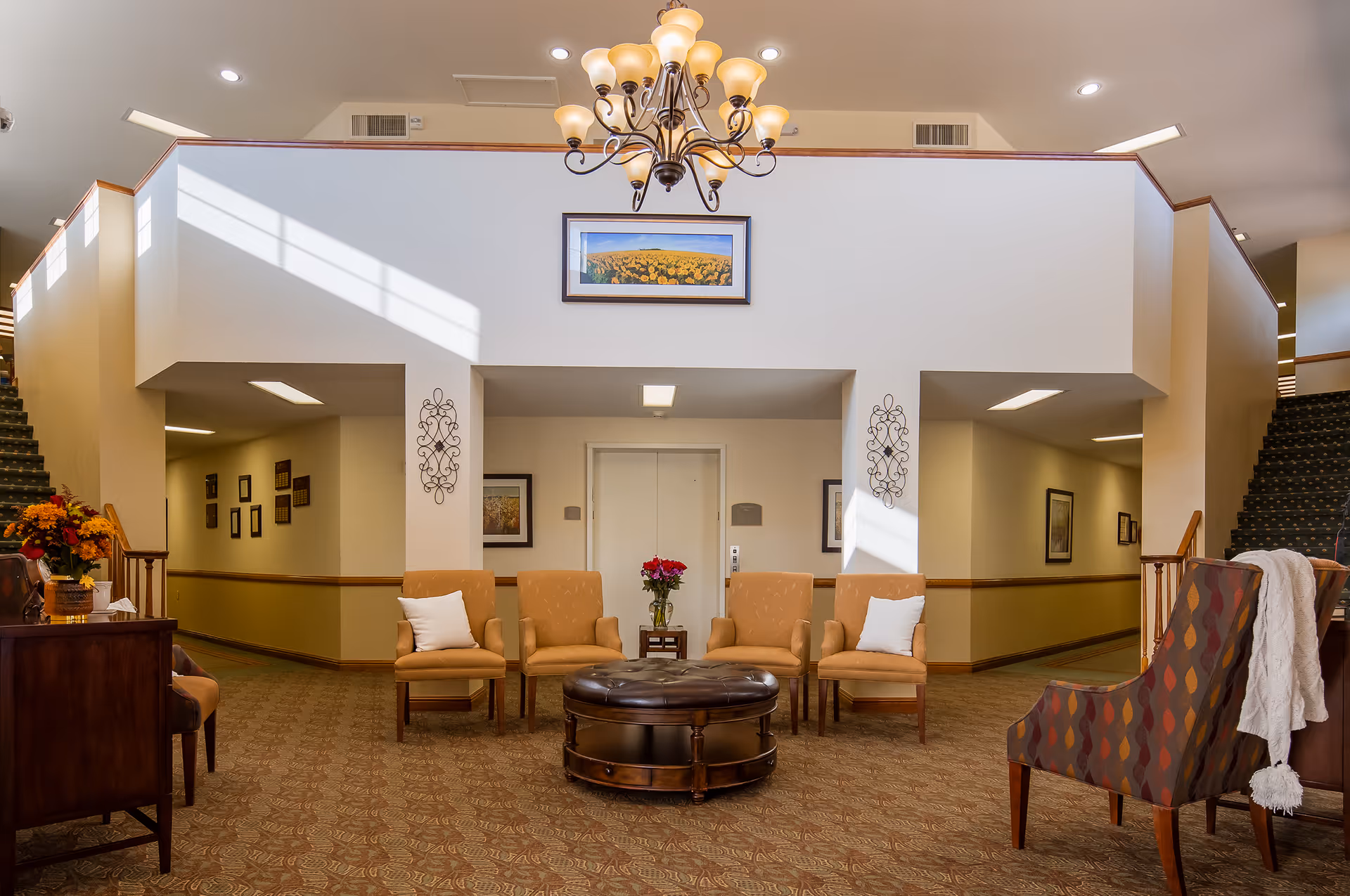 A spacious and well-lit common area in a senior living facility with four tan armchairs arranged around a round leather ottoman. There is a small table with a vase of red flowers in the center against a white wall with an elevator door. Two staircases with patterned carpet flank the seating area, and a chandelier hangs from the ceiling. The walls are decorated with framed pictures and metal wall art.