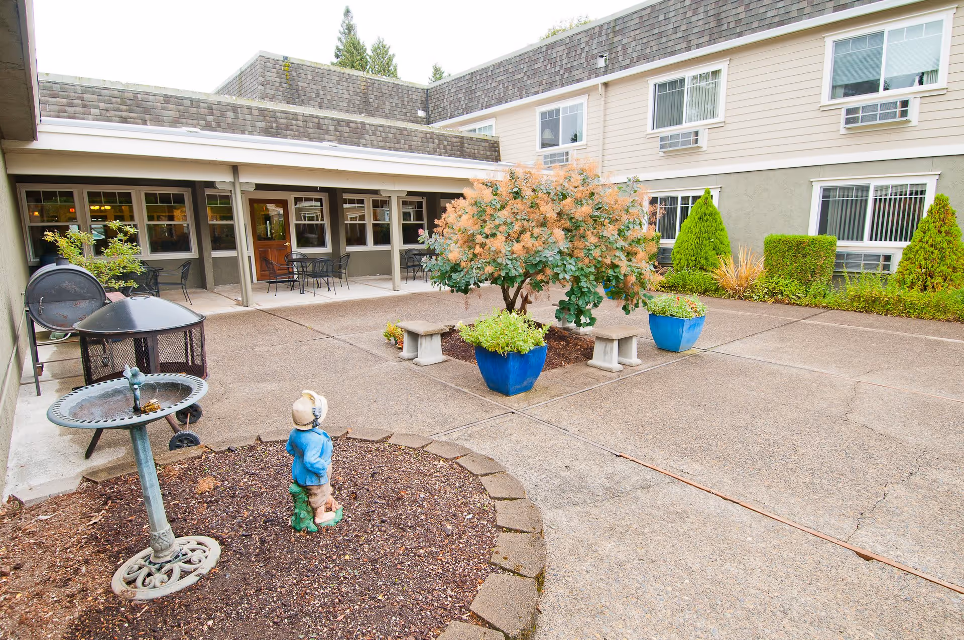 Outdoor courtyard area at Royalton Place featuring a paved patio with two concrete benches, two large blue planters with greenery, a small tree, a birdbath, a small garden statue of a child, and a covered seating area with tables and chairs adjacent to the building.