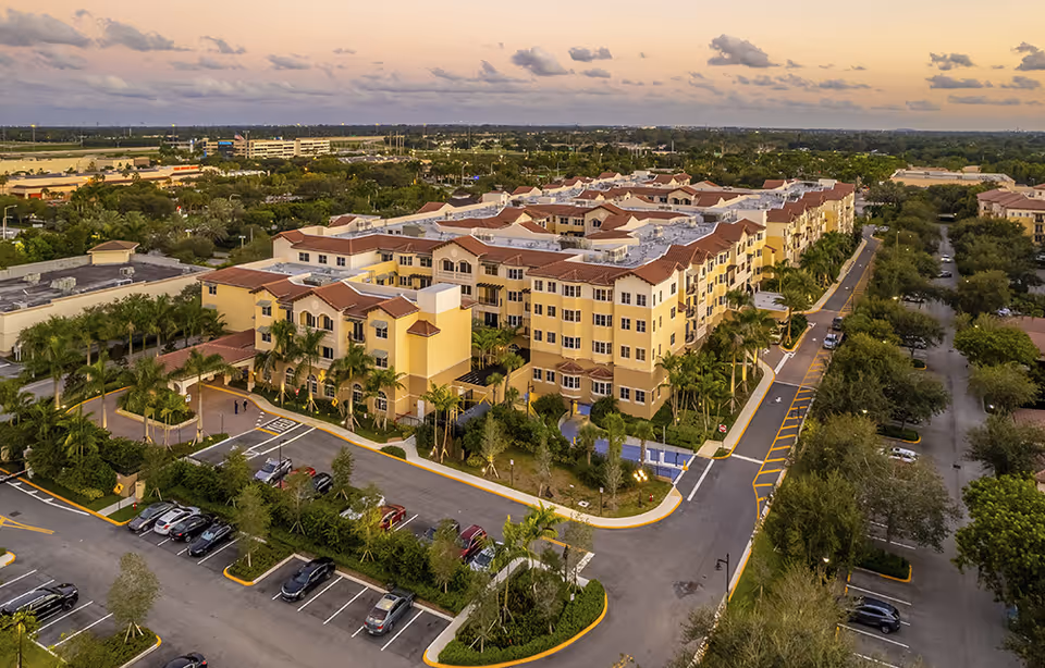 Aerial view of The Palace at Weston, a large senior living facility with multiple connected buildings featuring yellow walls and red-tiled roofs, surrounded by palm trees and parking lots under a cloudy sky at sunset.