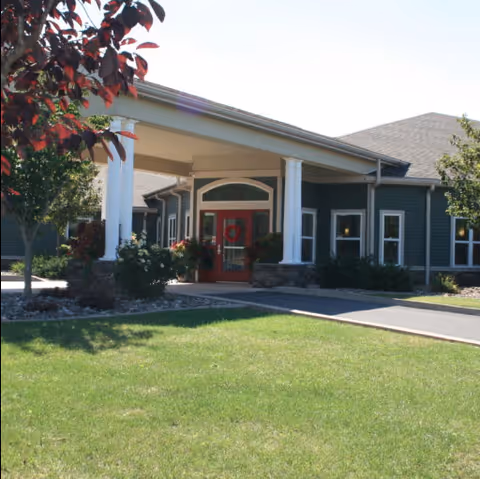 Exterior view of a single-story senior living community building with a covered entrance supported by white columns, a red door, and surrounding greenery including trees and bushes under a clear sky.
