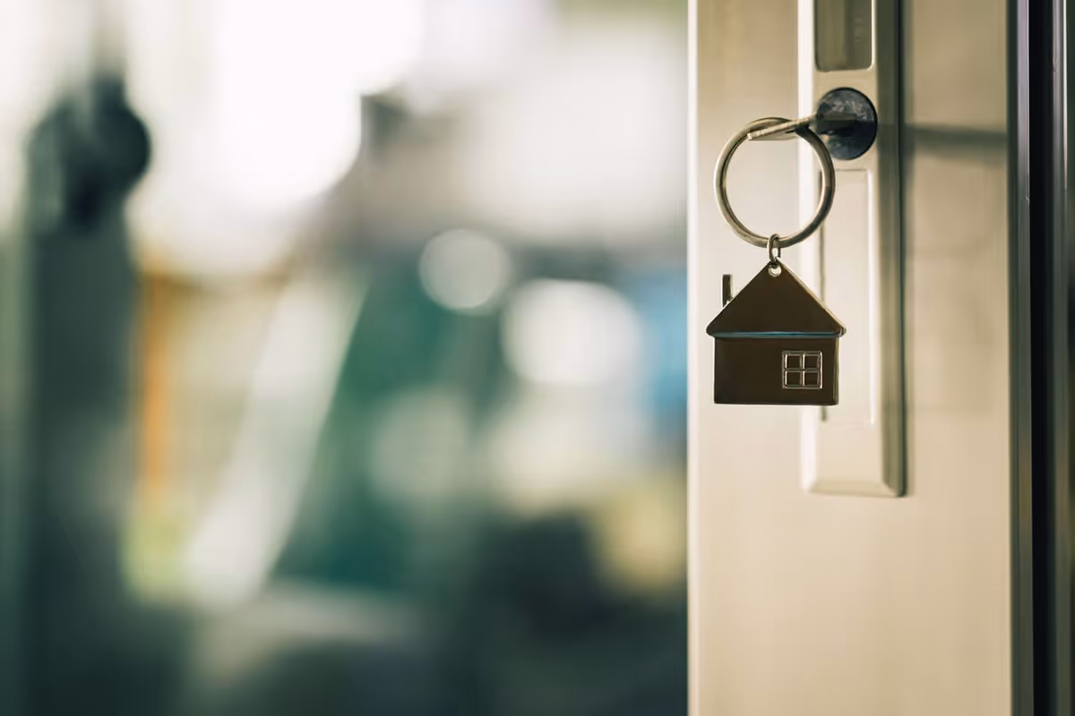 Close-up of a door key with a house-shaped keychain hanging in a lock against a blurred interior background.