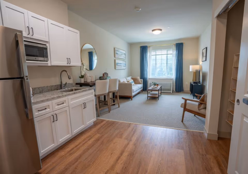 Open-plan apartment interior showing a kitchenette in the foreground and a furnished living area with a sofa and large window in the background.
