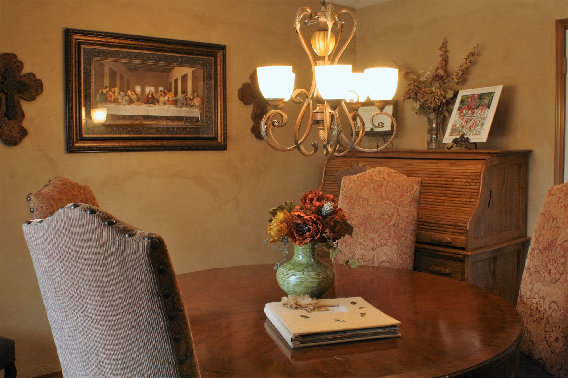 A warmly lit dining room with a round wooden table surrounded by upholstered chairs. A green vase with a bouquet of flowers sits on the table along with a closed book or album. A chandelier with five glowing lights hangs above the table. On the wall is a framed picture of The Last Supper and two decorative crosses. In the background, there is a wooden roll-top desk with a framed photo and a vase of dried flowers on top.