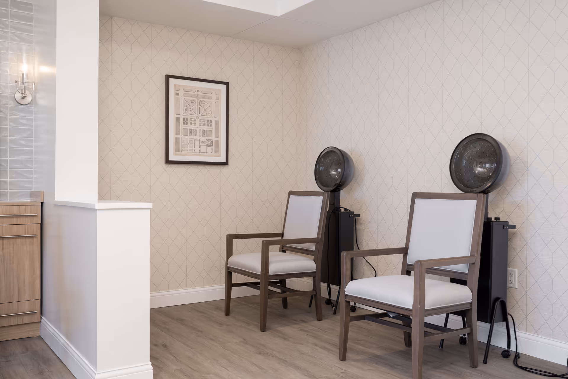 A small salon area with two wooden chairs featuring white cushions, each positioned in front of a black hair dryer hood. The walls have a light patterned wallpaper, and a framed map or artwork hangs on the wall. The floor is light wood, and part of a wooden cabinet and a wall sconce are visible on the left side.