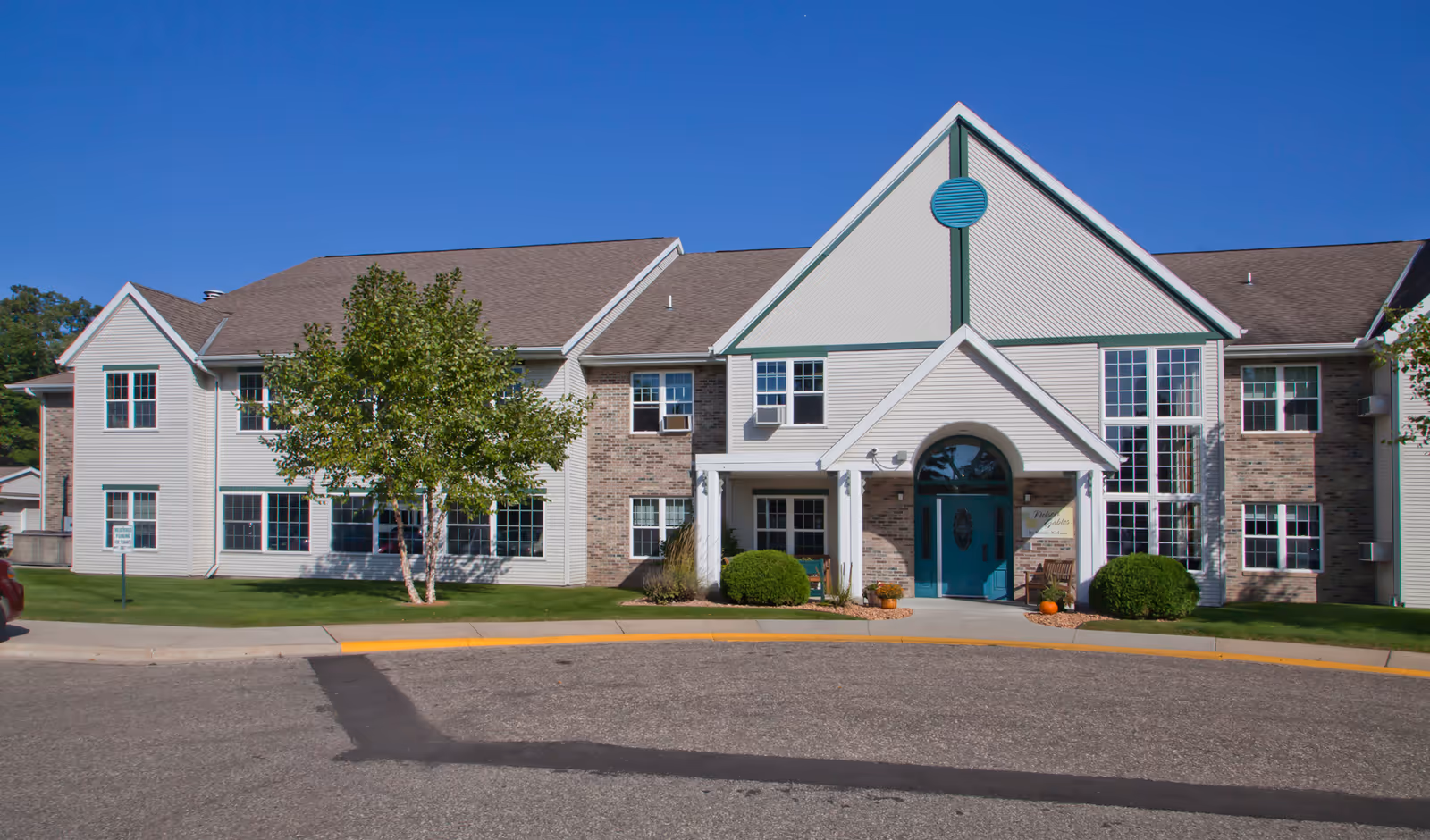 Front exterior view of a two-story senior living facility building with a peaked roof, large windows, a blue entrance door, and some landscaping including bushes and a tree. The sky is clear and blue.