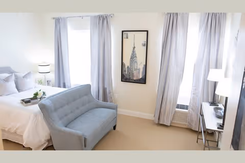 A bright bedroom featuring a neatly made bed with white linens and pillows, a small gray loveseat at the foot of the bed, two windows with light gray curtains, a framed picture of a cityscape on the wall, a bedside table with a lamp and small decor items, and a desk with a lamp near the window.