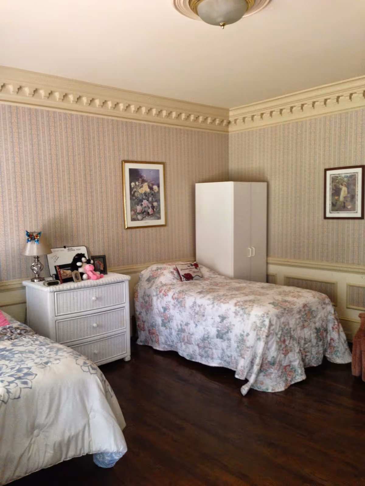 A cozy bedroom with two single beds covered in floral bedspreads. Between the beds is a white nightstand with a lamp, stuffed animals, and framed photos. The room has patterned wallpaper, crown molding, and two framed pictures on the walls. A white wardrobe is placed in the corner on a wooden floor.
