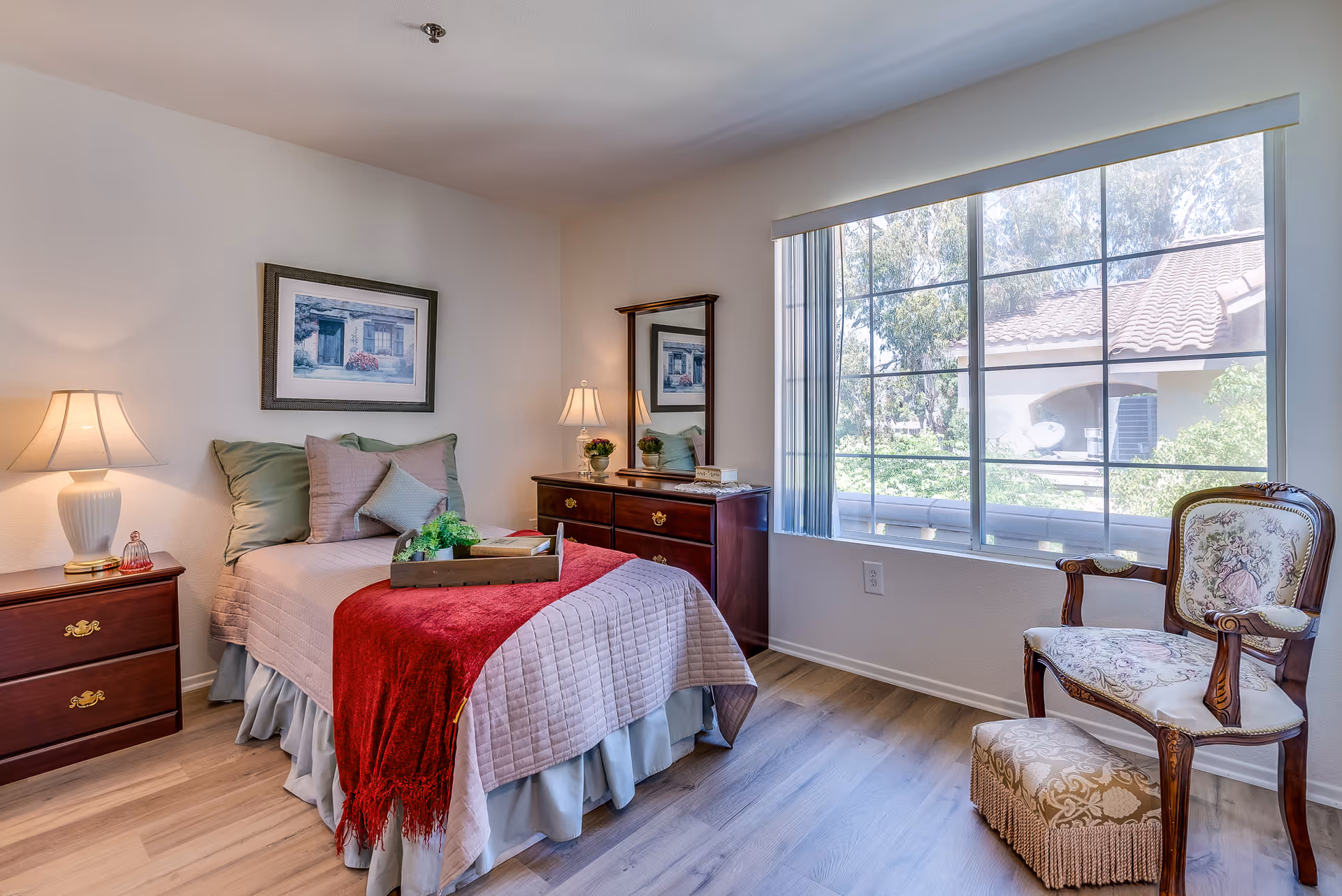 Well-lit single bedroom with a made bed, nightstands and dresser, a large window, and an upholstered chair.