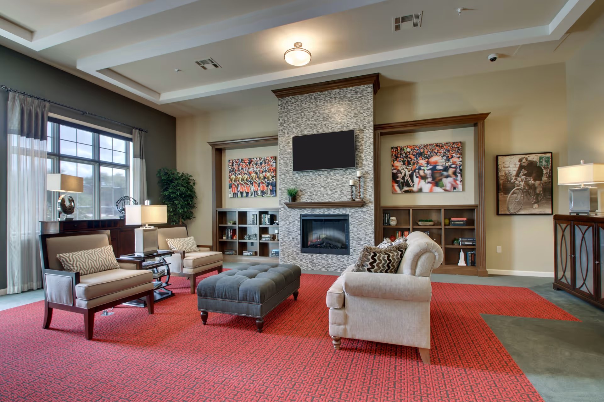 A cozy senior living facility common area with a red patterned carpet, beige armchairs, a gray tufted ottoman, and a beige sofa with patterned pillows. The room features a modern fireplace with a mounted flat-screen TV above it, built-in bookshelves with decorative items, large windows with curtains, and framed artwork on the walls. There are also table lamps and a potted plant adding to the warm ambiance.