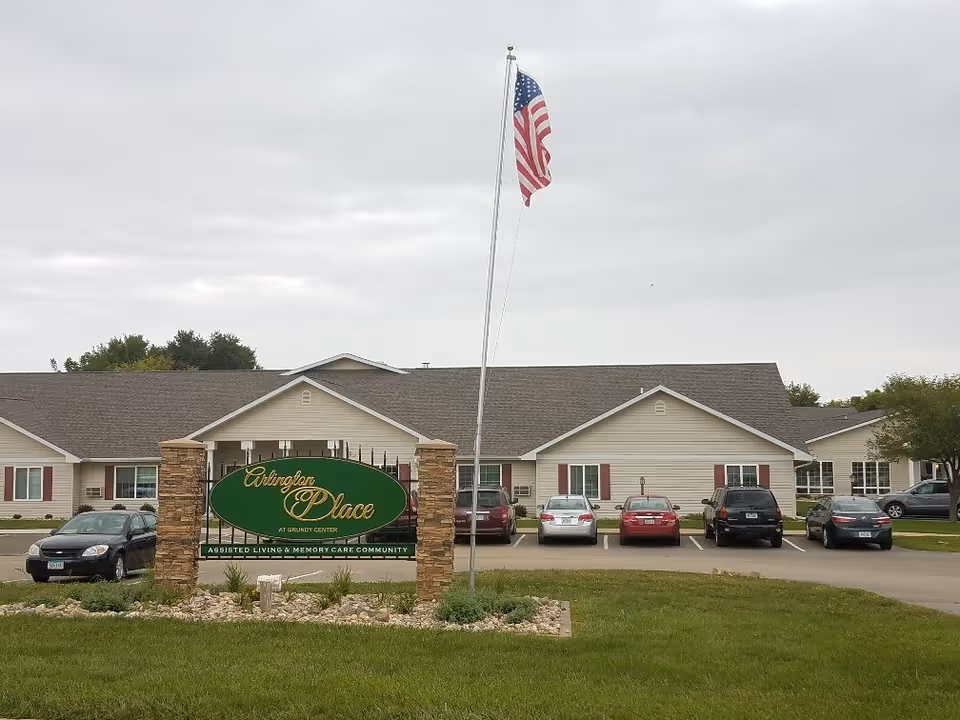 Exterior view of Arlington Place at Grundy Center, an assisted living and memory care community. The building is a single-story structure with beige siding and a gray roof. Several cars are parked in front of the building. A green sign with gold lettering is displayed on stone pillars near the entrance, and an American flag flies on a flagpole nearby under a cloudy sky.