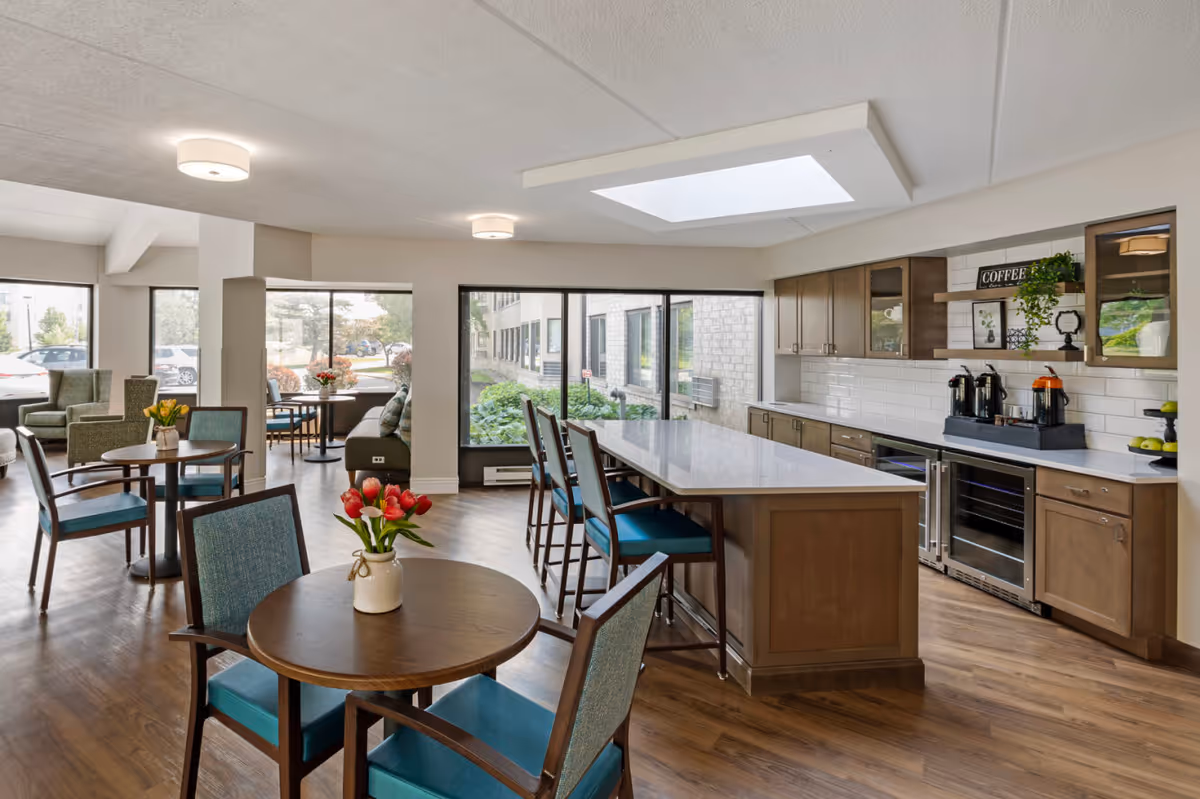 Bright communal dining area with round tables and teal-upholstered chairs, a large kitchen island, and windows overlooking landscaping.