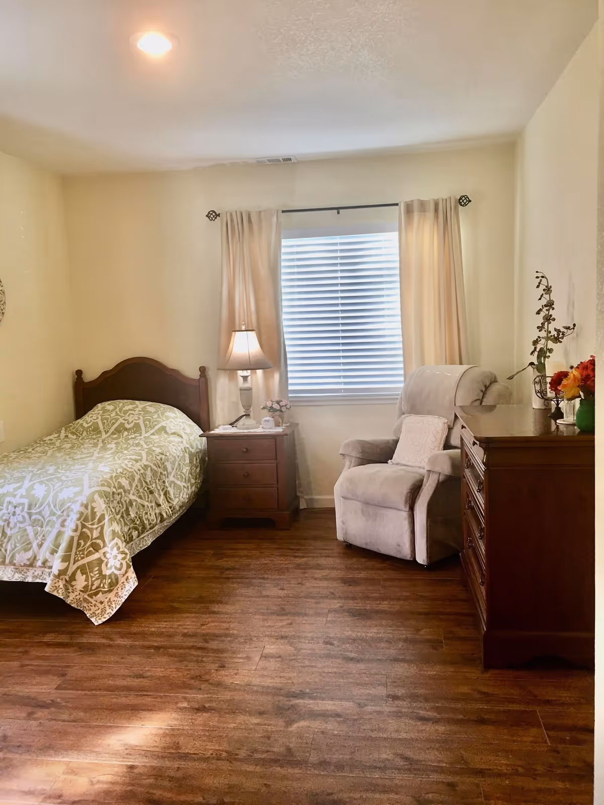 A cozy bedroom with a single bed covered in a green and white patterned bedspread, a wooden nightstand with a lamp and a phone, a comfortable beige armchair with a pillow, a wooden dresser with flowers in vases, and a window with closed blinds and beige curtains.