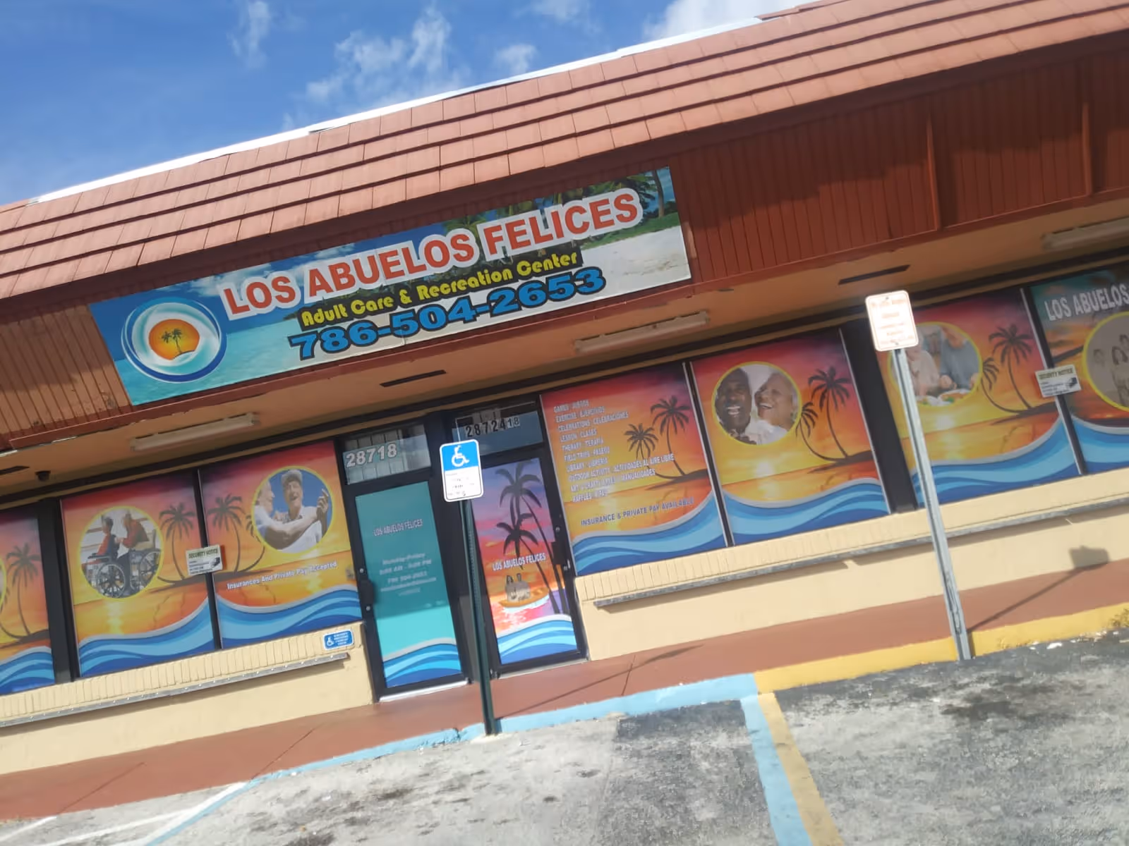 Exterior view of Los Abuelos Felices Adult Care & Recreation Center with colorful signage and window graphics depicting happy elderly people and palm trees. The building has a red tiled roof and a parking area with handicapped parking spaces in front.