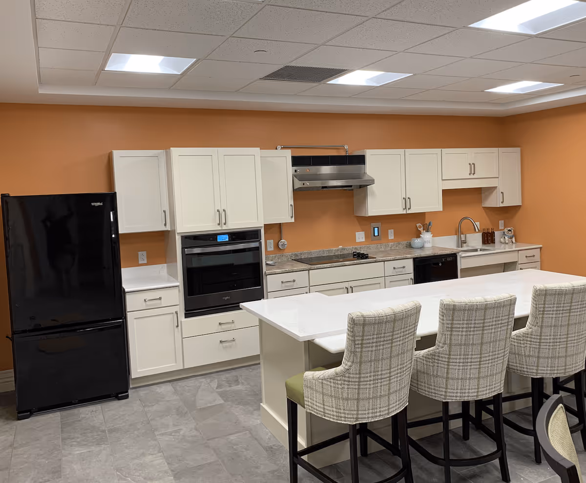 A modern kitchen with white cabinets, a black refrigerator, built-in oven, stovetop with a stainless steel range hood, and a white countertop island with four plaid upholstered bar stools. The walls are painted orange and the floor is tiled in gray.