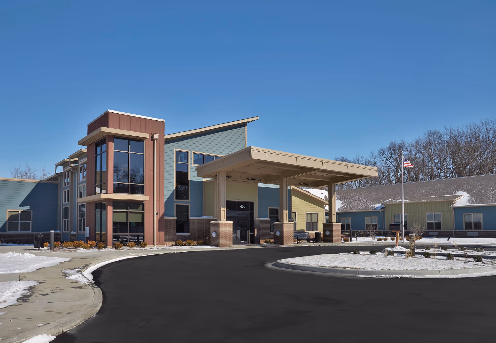 Front entrance of a senior living facility with a covered porte-cochere, circular driveway, and snow on the ground.