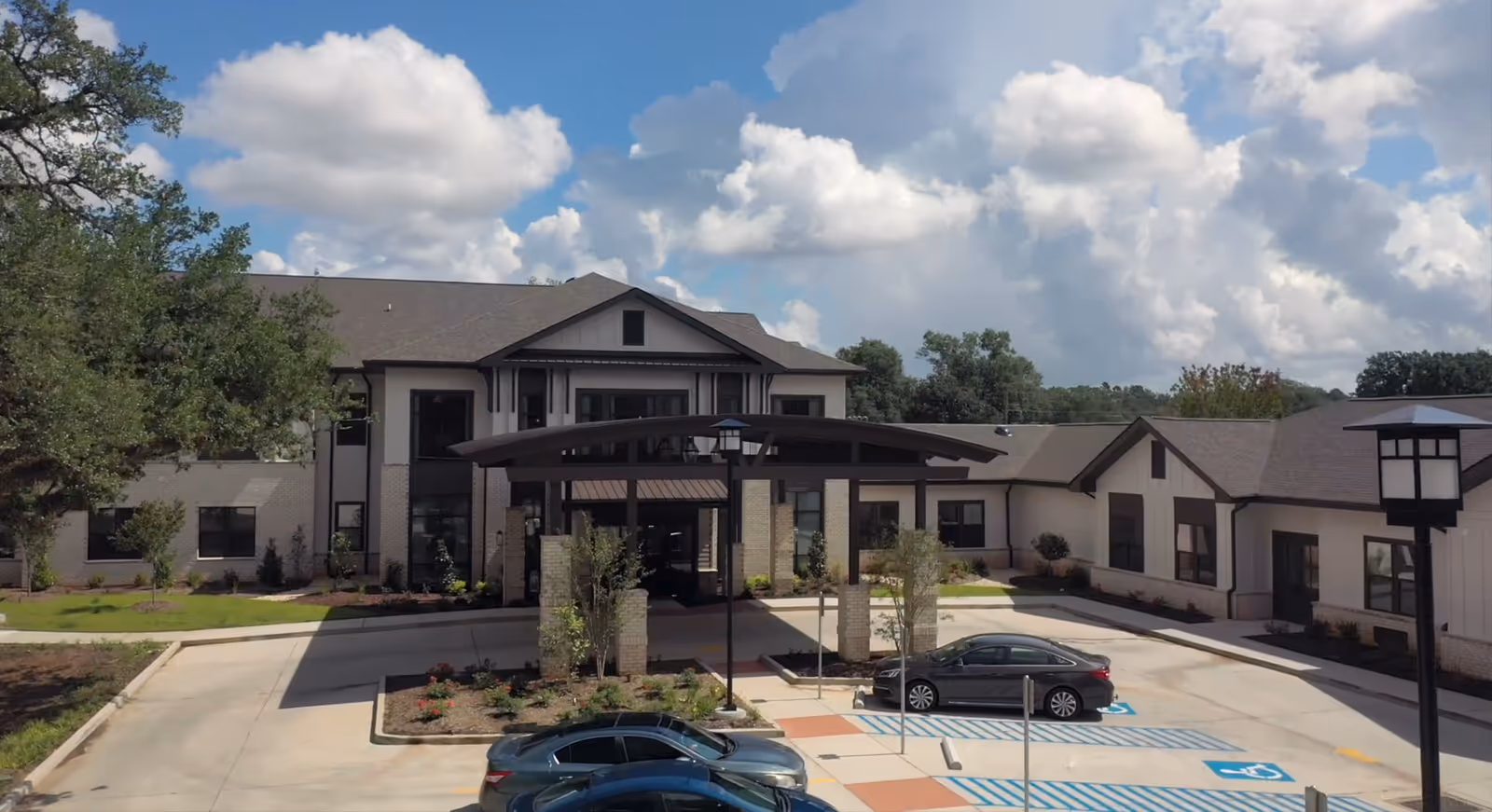 Front exterior of a senior living building with a covered entrance canopy, parking spaces and cars under a partly cloudy sky.