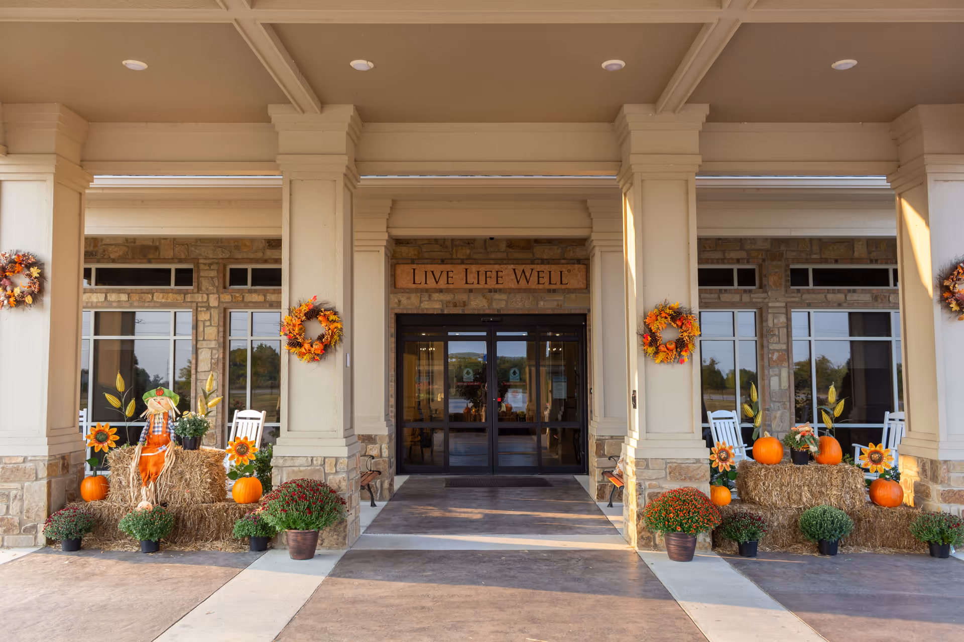 Front entrance of a senior living facility decorated for fall with pumpkins, hay bales and a 'Live Life Well' sign above the glass doors.