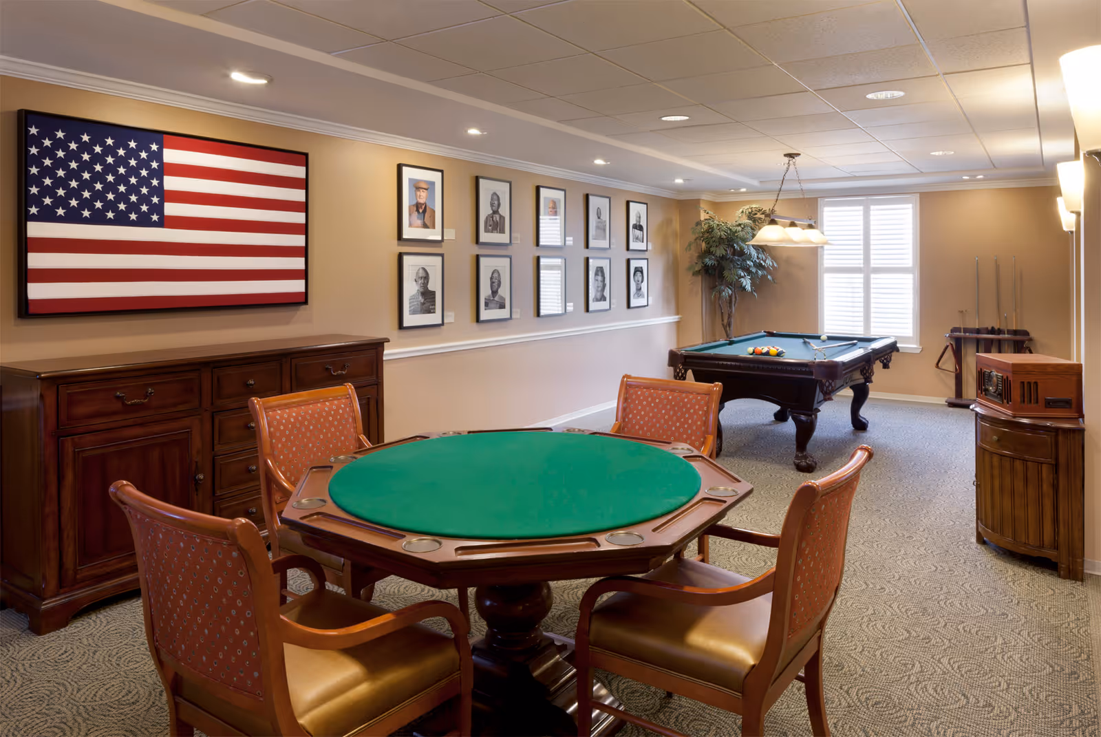 A senior living facility game room with a green felt card table surrounded by four wooden chairs with red cushioned seats. In the background, there is a pool table with billiard balls and cues, a large American flag framed on the wall, and several framed black and white portraits. The room has beige walls, carpeted floor, and a window with white shutters letting in natural light.