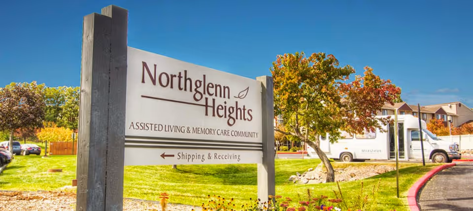 Outdoor view of a sign for Northglenn Heights Assisted Living & Memory Care Community, with a grassy area, trees with autumn foliage, parked cars, and a white van in the background under a clear blue sky.