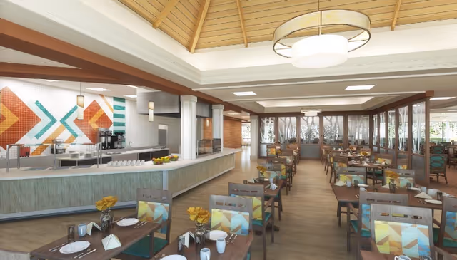 A spacious dining room with multiple wooden tables and chairs arranged neatly. Each table is set with plates, cups, napkins, and small vases containing yellow flowers. The room features large windows allowing natural light to enter, a high ceiling with wooden beams, and a modern light fixture. A serving counter with a colorful geometric tile backsplash is visible on the left side.