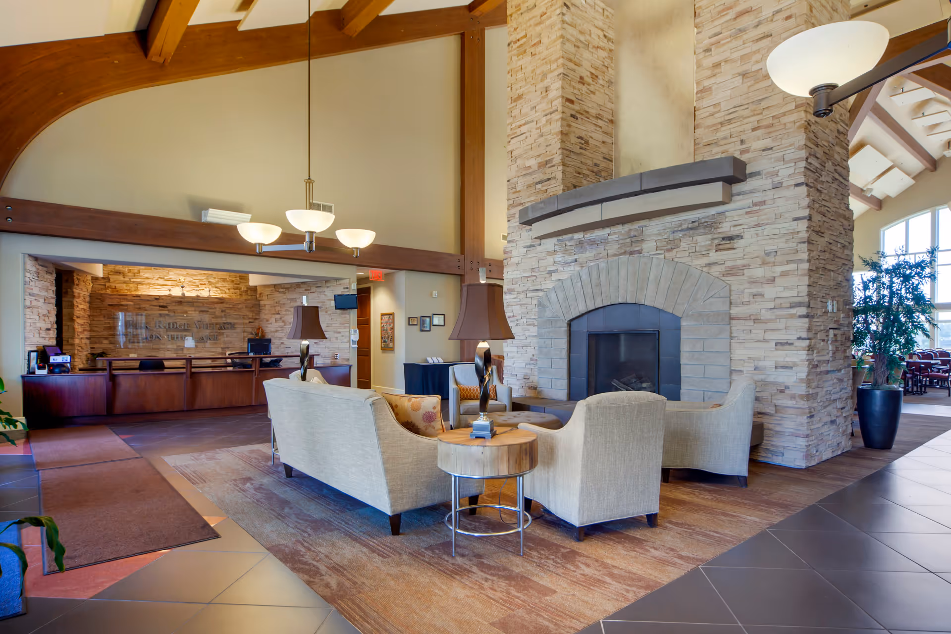 A spacious and warmly lit assisted living facility lobby with a large stone fireplace surrounded by beige armchairs and a sofa. There is a wooden side table with a lamp in the center, and a reception desk with a sign reading 'Elk Ridge Village on the Lake' in the background. The area features high ceilings with exposed wooden beams and large windows letting in natural light.