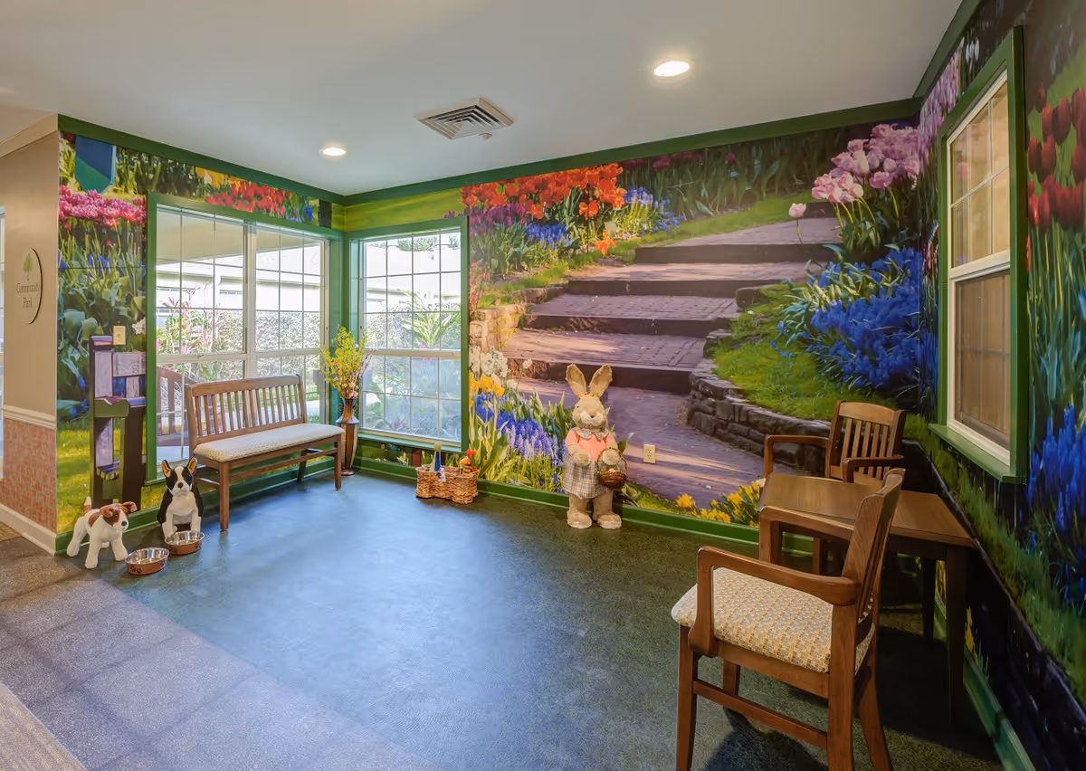 A cozy indoor seating area with a wooden bench and two wooden chairs around a small table. The walls are decorated with a colorful mural of a garden path surrounded by flowers. There are two large windows letting in natural light. Plush dog toys with bowls and a large stuffed bunny holding a basket add a playful touch to the space.
