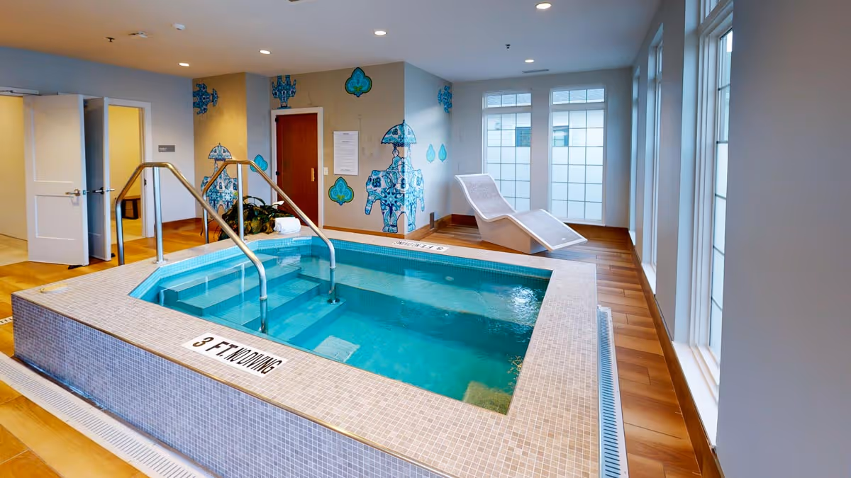 Indoor therapy pool with steps and handrails in a bright room featuring a lounge chair, decorative wall art, and large windows.