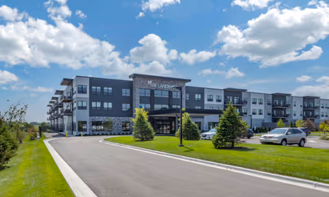 Three-story senior living building with a driveway, landscaped lawn, and parked cars under a blue sky.