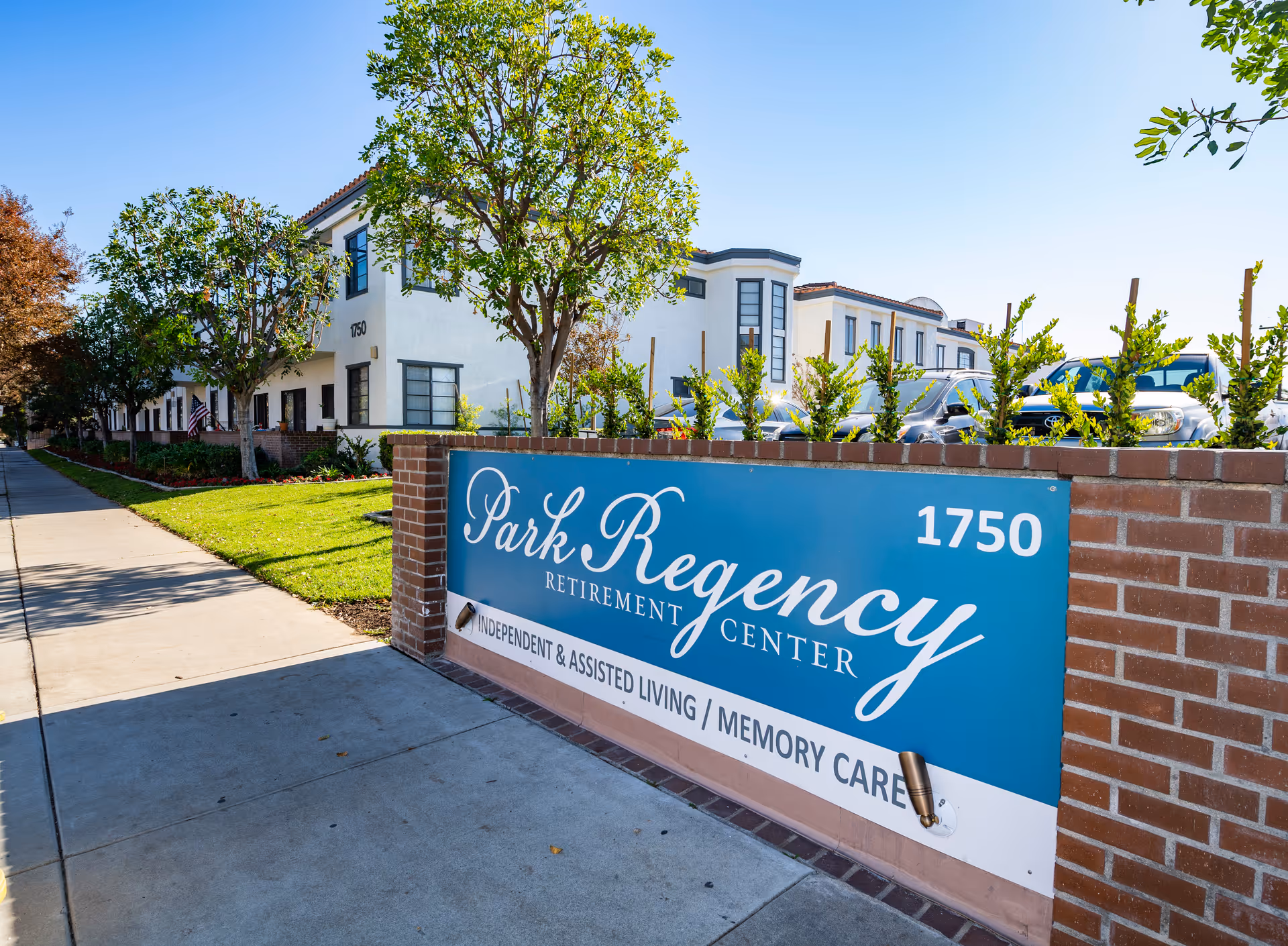 Exterior view of Park Regency Retirement Center with a blue sign displaying the facility name and services including Independent & Assisted Living and Memory Care. The building is white with multiple windows, surrounded by trees and a sidewalk.