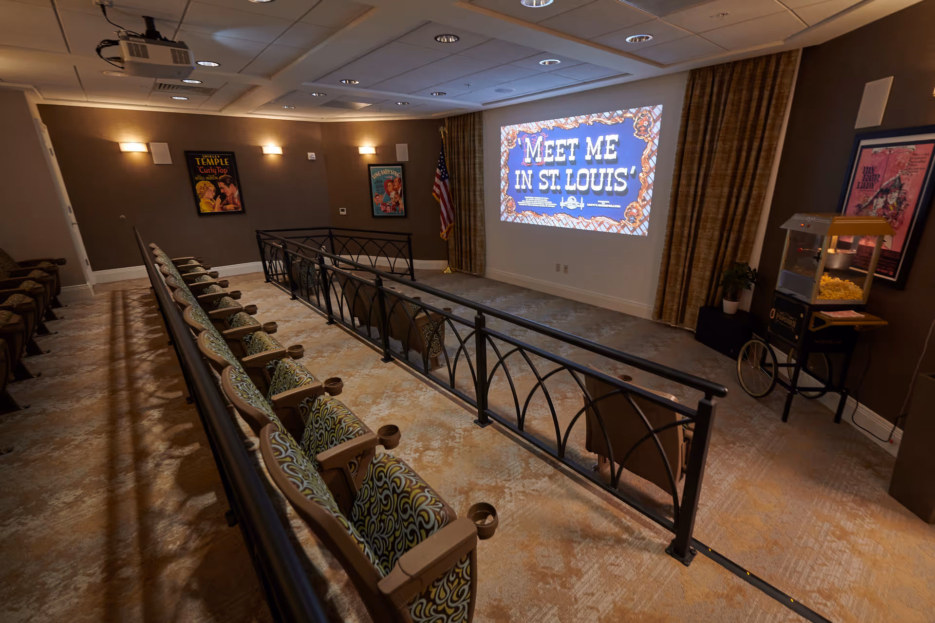 A small theater room with two rows of cushioned seats facing a large screen displaying the text 'MEET ME IN ST. LOUIS'. The room has brown walls with framed movie posters, an American flag in the corner, a popcorn machine on the right side, and ceiling lights. There is a black metal railing separating the seating area from the screen.