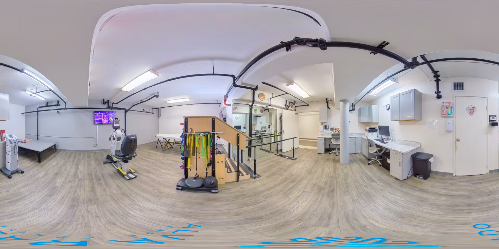 A spacious physical therapy room with light wood flooring and white walls. The room contains exercise equipment including a stationary bike, parallel bars, a set of stairs with handrails, resistance bands, and therapy balls. There is a desk area with a computer and office chair on the right side, and a TV mounted on the wall on the left. The ceiling has exposed black pipes and fluorescent lighting.