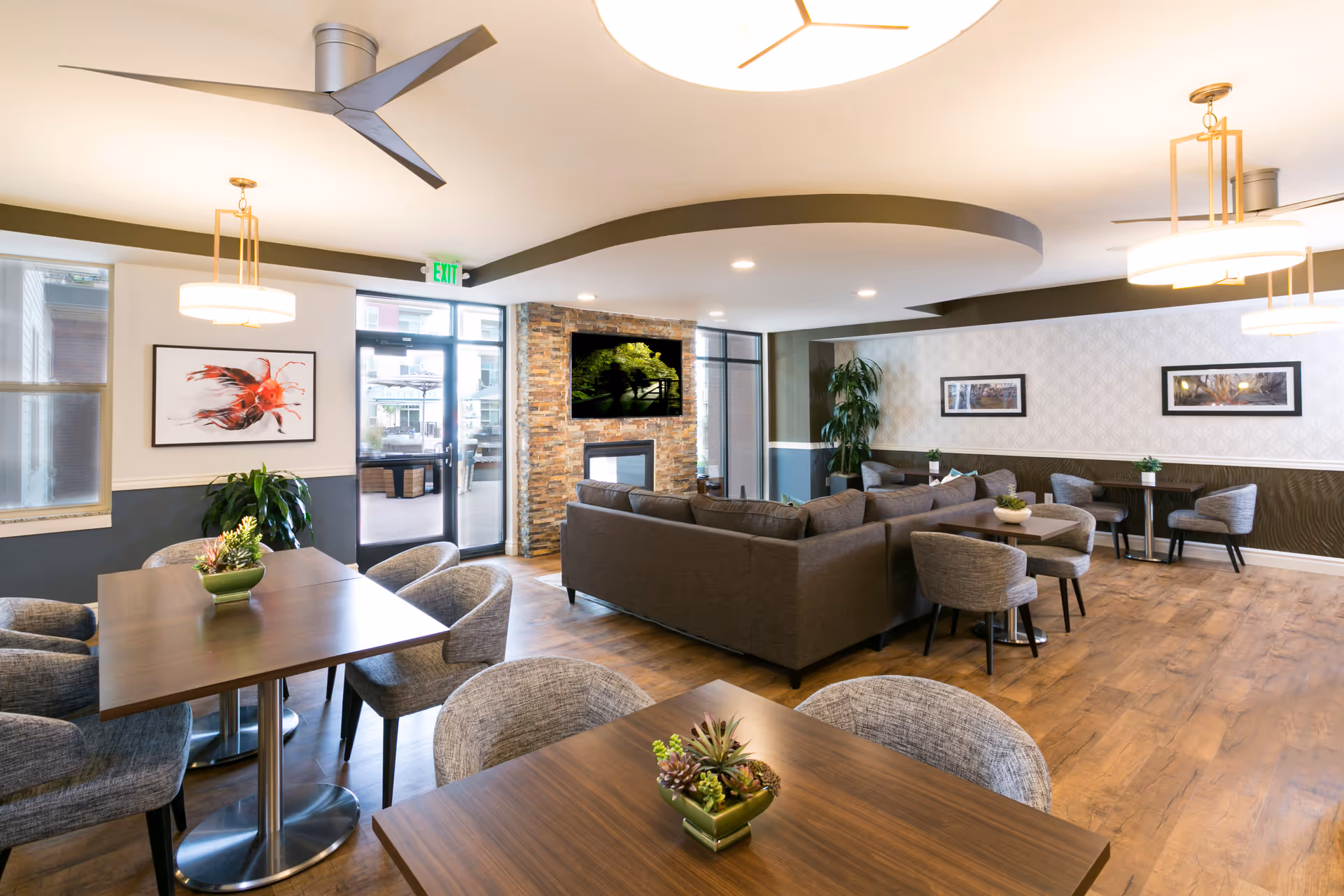 Spacious senior living common room with dining tables and upholstered chairs, a lounge area with sofas around a stone fireplace, and modern lighting.