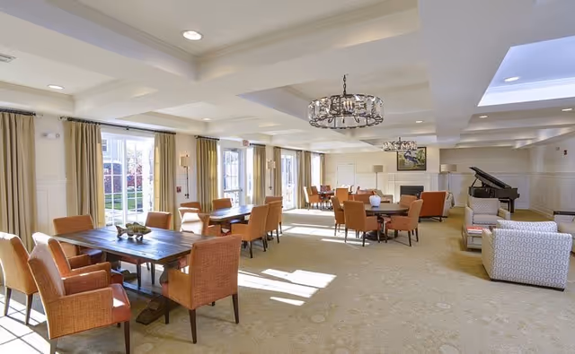 A spacious, well-lit common area with multiple wooden tables surrounded by orange upholstered chairs. Large windows with beige curtains allow natural light to fill the room. There is a chandelier hanging from the ceiling, a fireplace with a painting above it, and a grand piano in the corner. Comfortable armchairs are arranged near the piano.