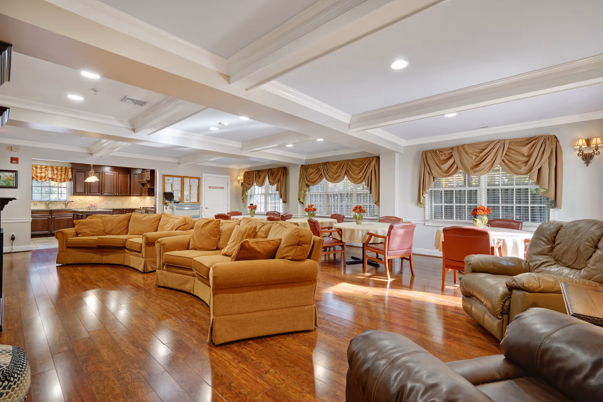 A spacious and well-lit common area in a senior living facility featuring comfortable tan sofas and leather armchairs arranged on polished wooden floors. In the background, there are round tables with red chairs and floral centerpieces near large windows with beige drapes. A kitchen area with dark wood cabinets and a tiled backsplash is visible in the far left corner.