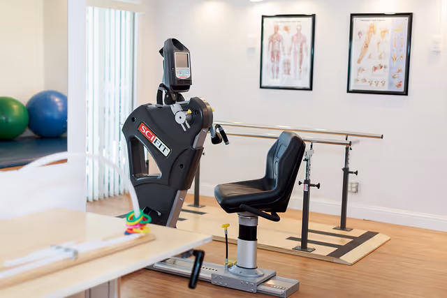 A physical therapy room with exercise equipment including a seated exercise machine labeled 'SCIFIT', parallel bars for walking support, anatomical charts on the wall, and exercise balls in the background.