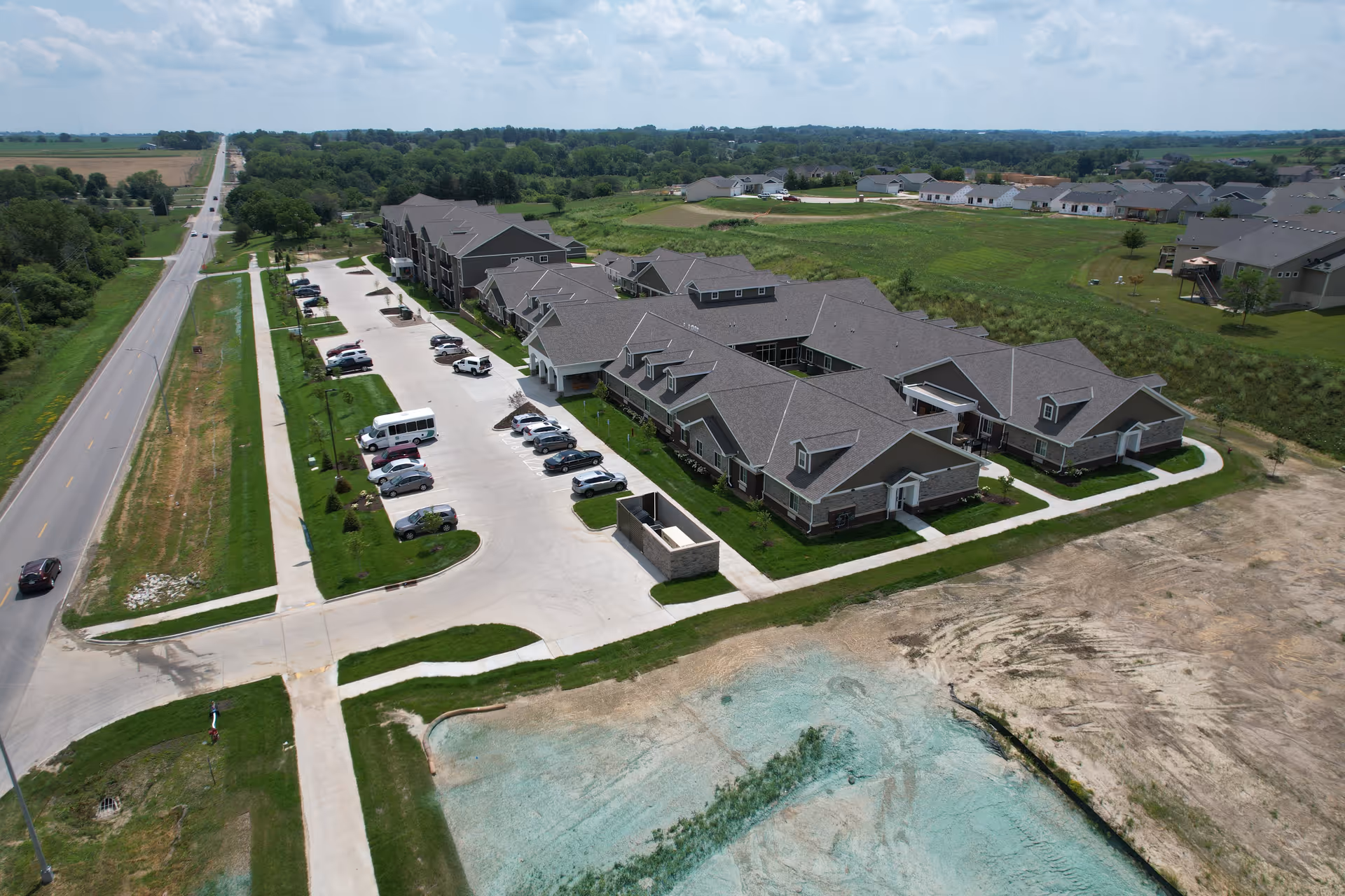 Aerial view of Edencrest at The Tuscany senior living facility showing multiple connected buildings with gray roofs, a parking lot with several cars and a shuttle bus, surrounded by green fields and a road on the left side.