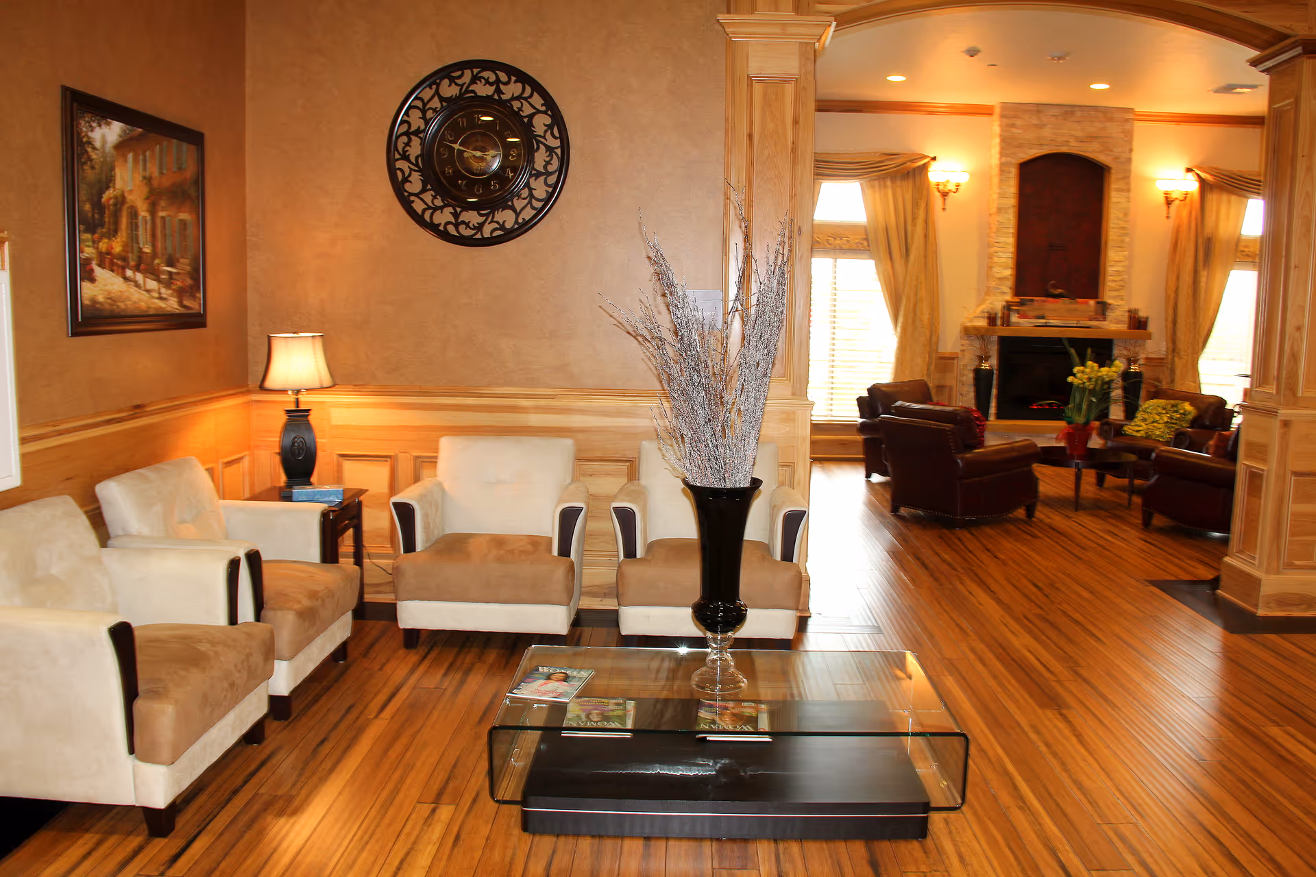 A warm, wood-accented common living room with upholstered armchairs, a glass coffee table and a fireplace in the background.