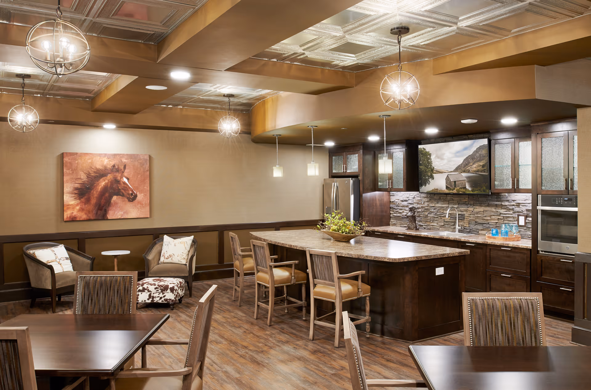 Communal kitchen and dining area with a large island, pendant lights, dining tables, and lounge seating in a senior living facility.