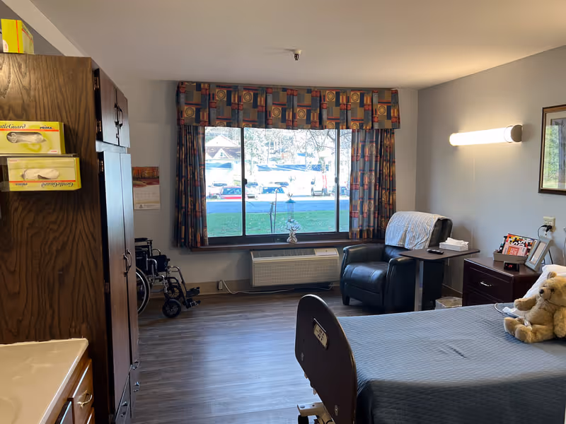 A senior living resident room with a bed, recliner, wheelchair, window with patterned valance, and wooden storage cabinet.