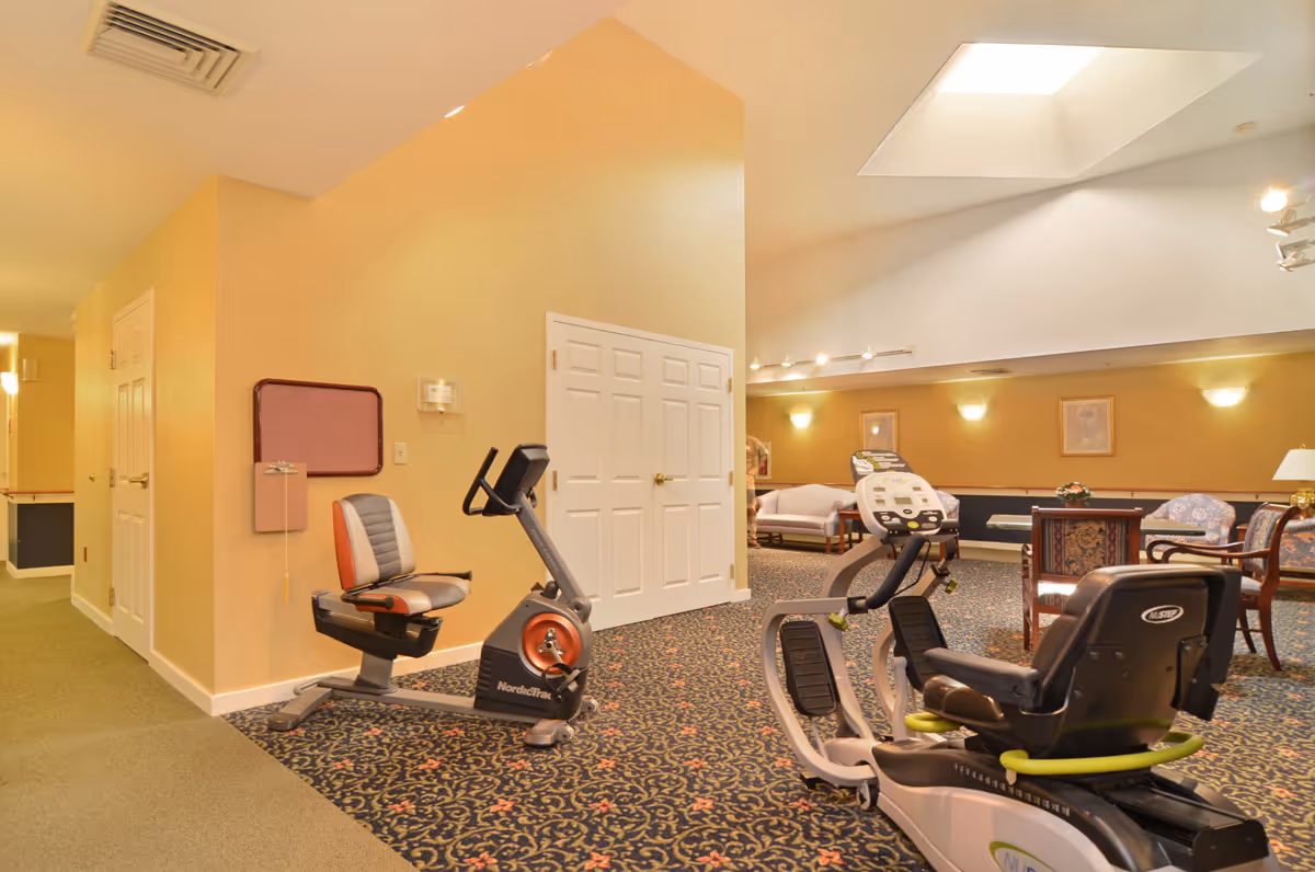 Interior view of a senior living facility room with exercise equipment including a recumbent bike and a rowing machine. The room has patterned carpet, beige walls, a skylight, and seating areas with chairs and tables in the background.