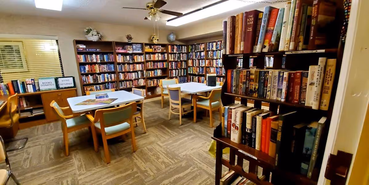 Community library room with bookshelves lining the walls, tables and chairs, and a book donation/return area.