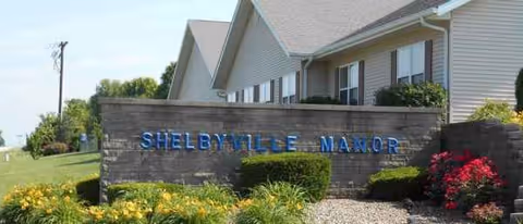Shelbyville Manor sign on a stone wall with a beige building and landscaped grounds.