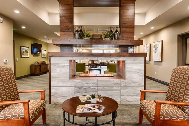 A cozy interior common area with two patterned armchairs facing a modern double-sided fireplace with a wooden mantel. A round wooden coffee table with magazines and a small plant is placed between the chairs. In the background, there are more seating areas and framed artwork on the walls.