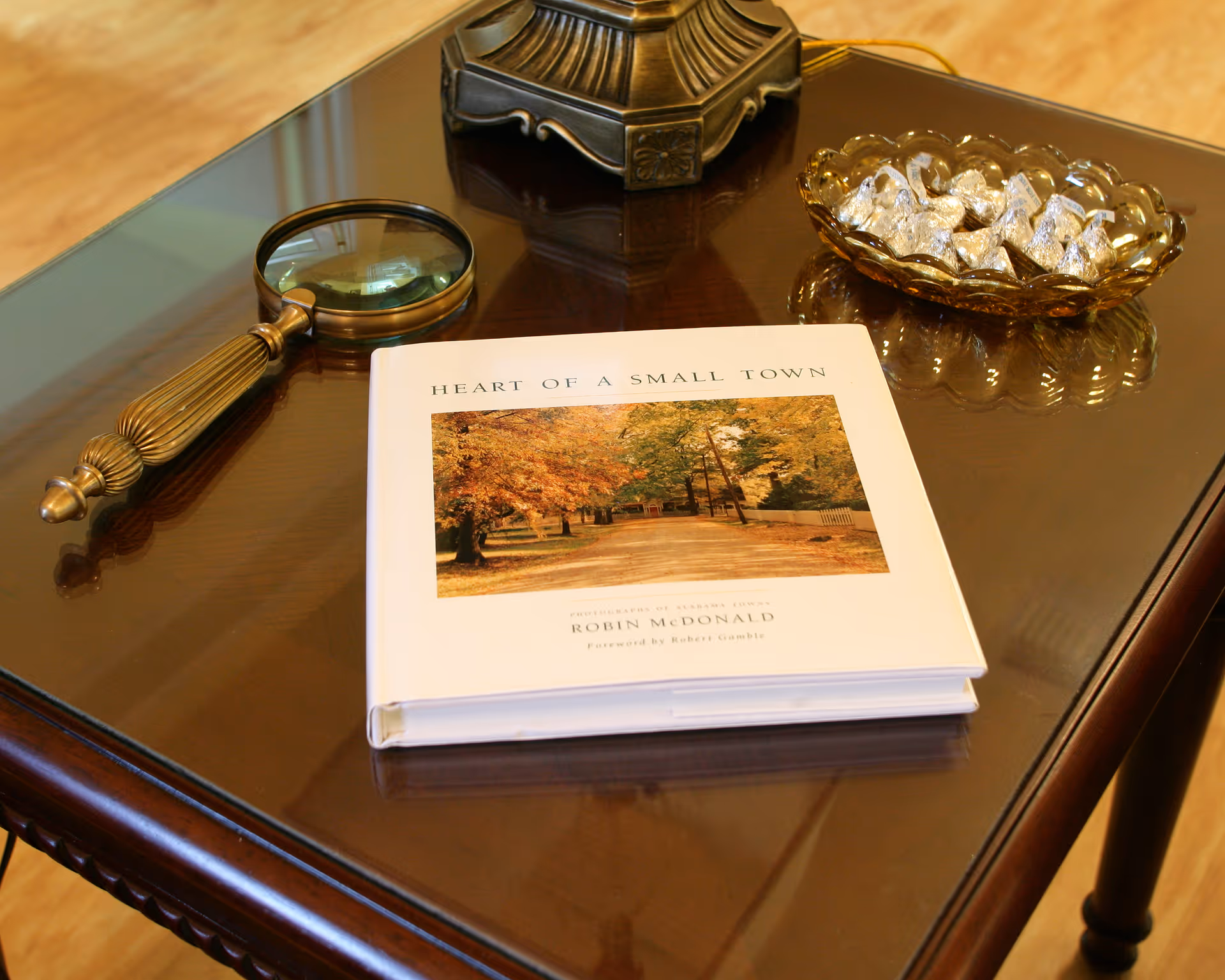 A close-up view of a polished wooden table with a magnifying glass, a decorative bowl filled with wrapped chocolates, and a book titled 'Heart of a Small Town' by Robin McDonald placed on it.