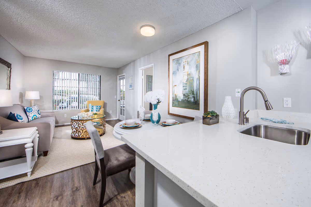 A bright and modern living area with a gray sofa adorned with blue and white patterned pillows, a yellow armchair, and a round glass coffee table with decorative items. The room features a large window with vertical blinds, a white side table with a lamp, and a kitchen counter with a sink and two place settings. A large framed abstract painting hangs on the wall.