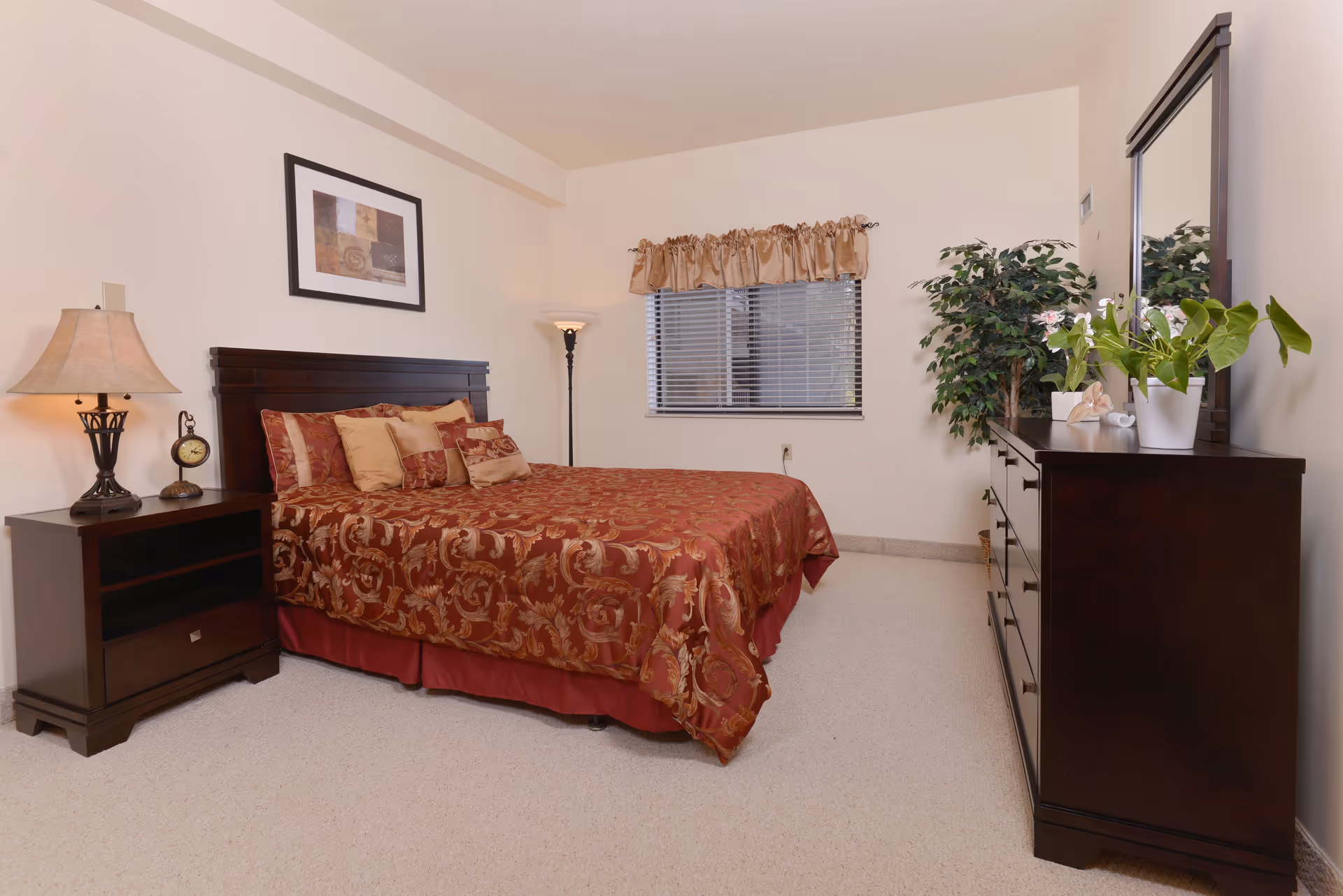 A bedroom with a red patterned bedspread, a nightstand and lamp to the left, and a dresser with mirror and plants to the right.