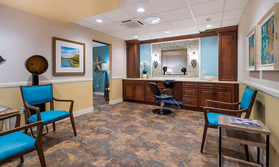 A well-lit salon area in a senior living facility featuring a styling chair in front of a large mirror with wooden cabinetry below. The room has a tiled floor, light-colored walls with a blue accent wall, and several turquoise cushioned chairs along the walls. There are framed artworks on the walls and a hair dryer hood in the corner.