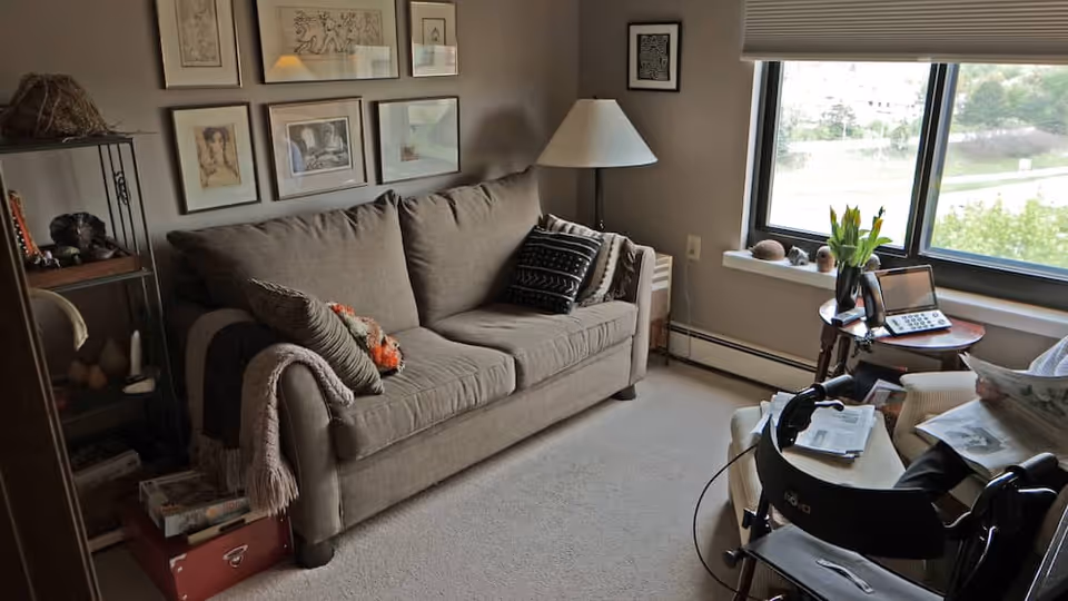 Cozy living room with a beige sofa under framed artwork, a floor lamp, a windowed side table with a phone and flowers, and a walker beside an armchair.